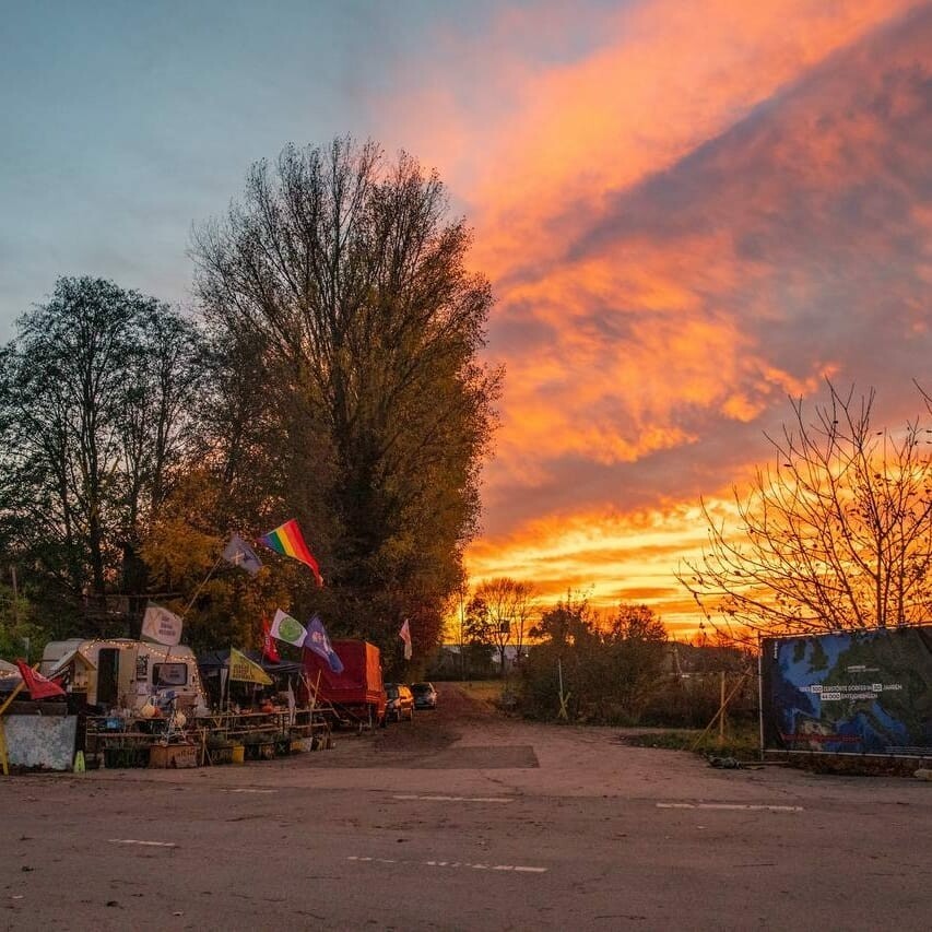 A sunset photo of the entrance to Lützerath.  To one side one can see a white trailer, full of positive flags, along with other camping structures.