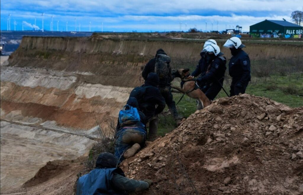 A photo right at the edge of the enourmous mine pit.  Cops in riot gear send an attack dog on protesters who are centimetres from the edge.