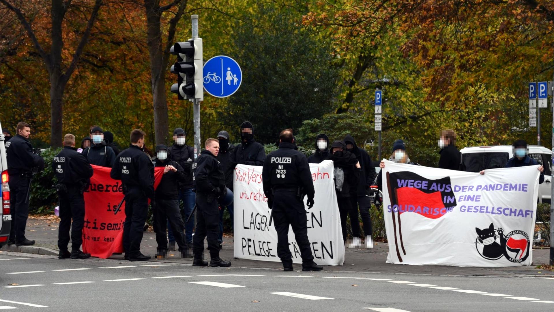 Abgebildet ist eine Gruppe von Antifaschist:innen, die an einer Kreuzung mehrere Transparente halten. Darauf steht zum Beispiel der Text "Querdenken ist antisemitisch". Vor der Gruppe stehen aufgerüstete Polizist:innen mit Schlagstöcken und Demoausrüstung.