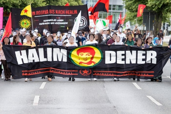 Das Foto zeigt eine Demonstration auf der Straße. Vorne ist ein großes Transparent mit der Aufschrift "Halim Dener" und dem Gesicht von Halim Dener. Im Hintergrund ist ein Transparent mit der Aufschrift "Von Rojava bis nach Berlin -  Kommunales Leben aufbauen und verteidigen". Es sind außerdem Flaggen zu sehen, von denen eine als YPG Fahne erkennbar ist. Einige Menschen tragen das Gesicht von Halim Dener als Maske. 