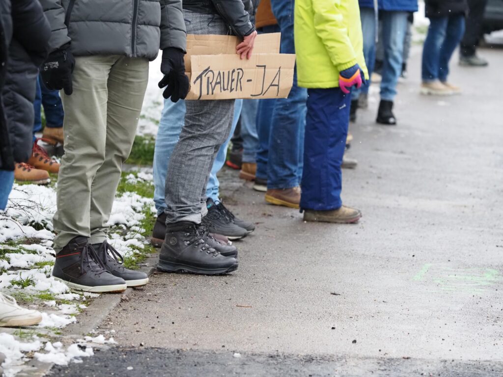 Menschen stehen auf der gegenüberliegenden Straßenseite. Eins trägt ein Schild, auf dem steht "Trauer -Ja"