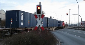Foto zeigt den ersten Uranerzkonzentrat beladenen Container auf dem zweiachsigen Containertragwagen und die vier mit Uranerzkonzentrat beladenen Container auf dem sechsachsigen Gelenk-Containertragwagen bei dem unbeschrankten Bahnübergang der Straße Veddeler Damm im Hamburger Hafen in Fahrtrichtung des Hafenbahnhof Hamburg-Süd
