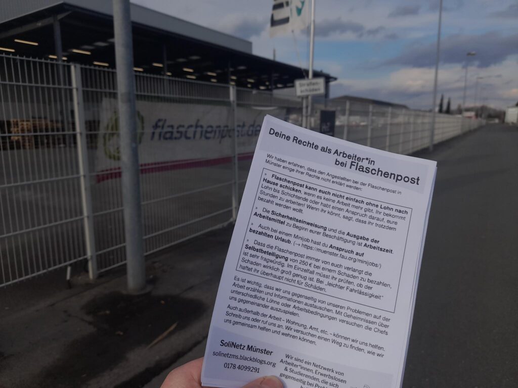 Auf dem Bild ist ein Flyer mit der Überschrift "Deine Rechte als Arbeiter:in bei Flaschenpost" zu sehen. Im Hintergrund ist die Flaschenpostniederlassung Münster erkennbar