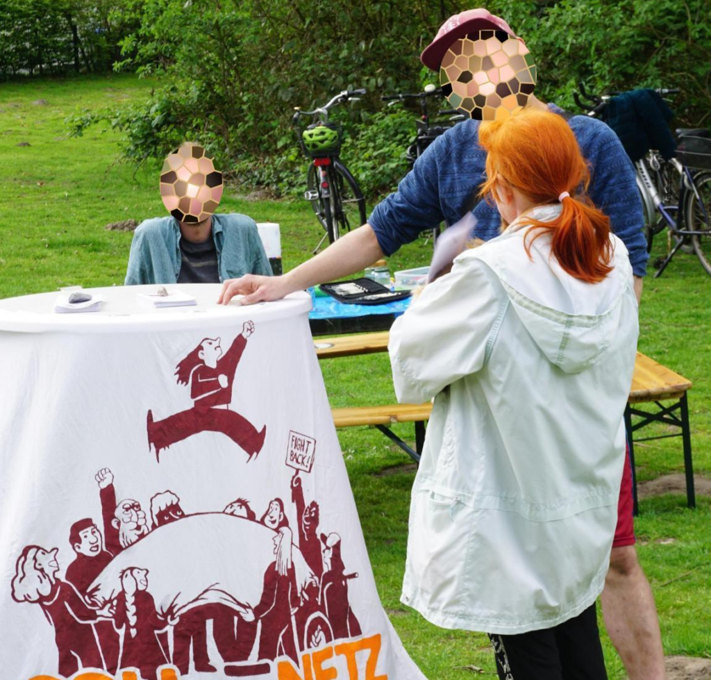 Auf dem Bild ist ein Stehtisch zu sehen, an dem ein Transparent mit dem Solinetz-Logo befestigt ist. An dem Tisch stehen zwei Personen und unterhalten sich, im Hintergrund sitzt eine weitere Person auf einer Bank an einem Tisch.