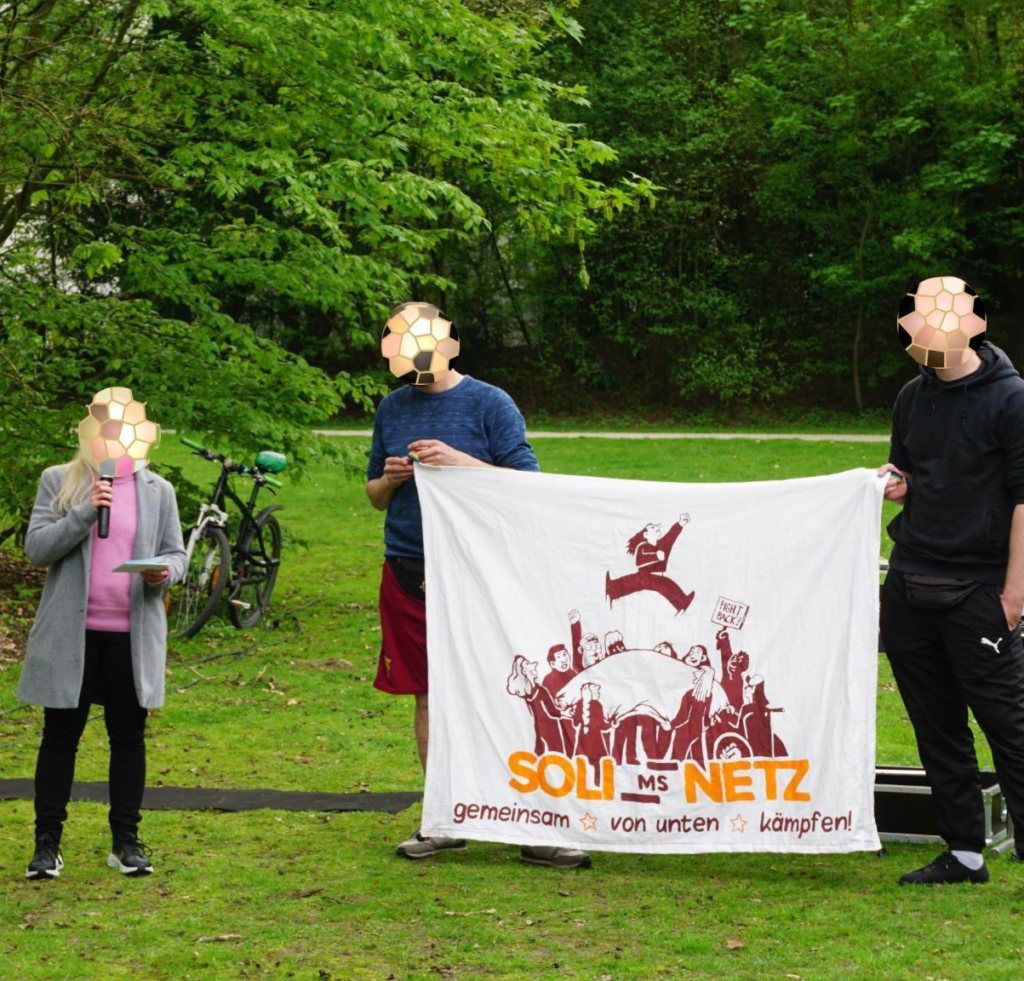 Auf dem Bild stehen drei Personen auf einer Wiese. Eine Person hat ein Mikrofon in der Hand und liest einen Text vor, die anderen beiden halten ein Transparent mit dem Solinetz-Logo.