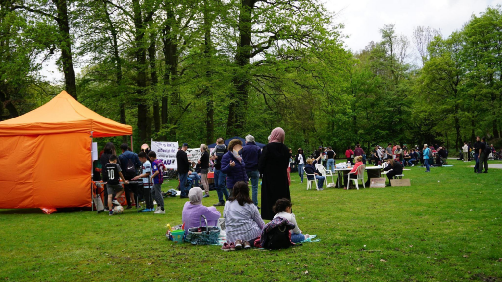 Auf dem Bild ist ein Park mit vielen Menschen zu sehen. Einige sind am Picknicken, einige stehen an einem orangenen Infopavillon und andere sitzen auf weißen Plastikstühlen.