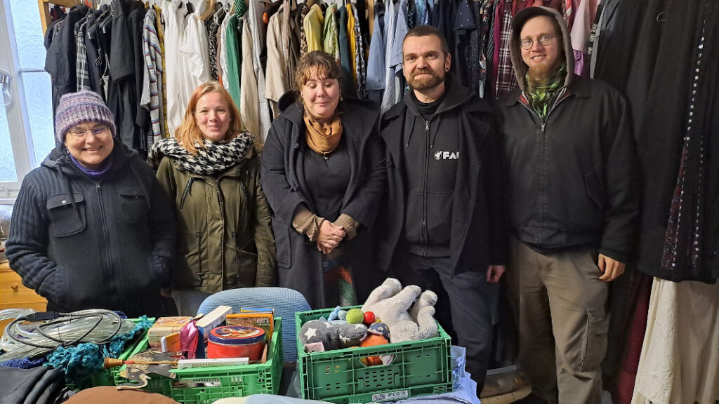 Personen vom Umsonstladen Nürtingen und Kollmarsreute angeordnet nach größe von klein bis groß stehen vor Kleidung auf Kleiderbügel und im Vordergrund sind Kisten für den Weihnachtsmarkt