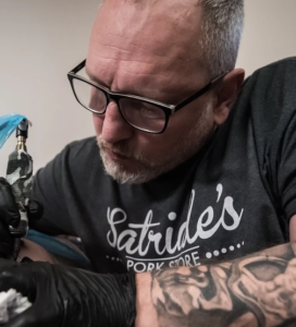Ein Mann Mitte 50 mit dunkler Brille, schwarzem T-Shirt und Schwarzen Gummihandschuhen beugt sich über etwas. Sein linker Arm ist tättowiert, in der anderen hält er eine Tättowiernadel