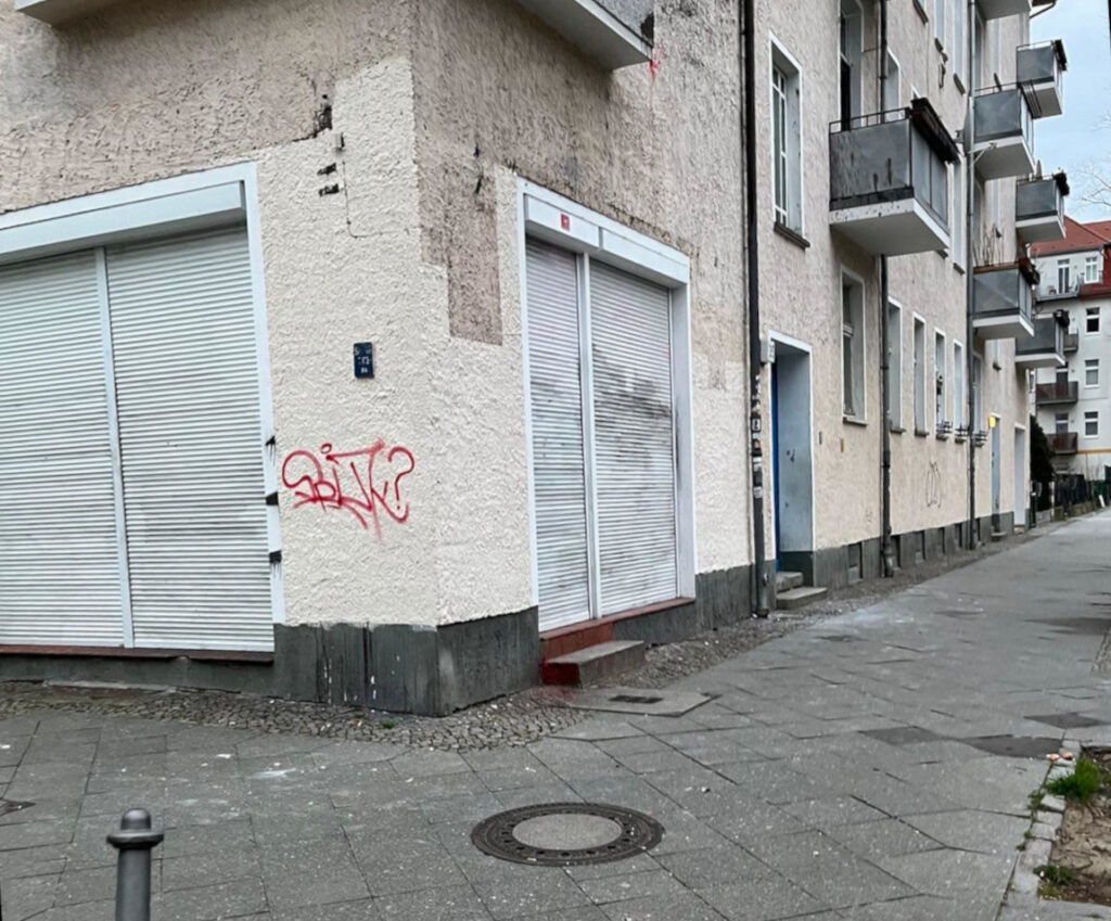 Ladengeschäft an einer Häuserecke. Die Jalousien sind heruntergelassen und ziemlich verschmutzt. Die Fassade hat unregelmäßige Abschnitte hellerer und dunklerer Farbe, darauf ist ein einzelnes rotes Graffiti.