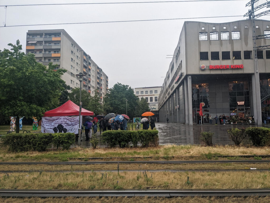 Der Platz vor dem Einkaufszentrum "Lindencenter" an einem regnerischem Tag. Hier steht ein Pavillion, daran hängt ein Transparent auf dem "schaut nicht weg - zusammen gegen Rassismus" auf vielen verschiedenen Sprachen steht. Daneben sind einige Menschen mit bunten Regenschirmen zu sehen